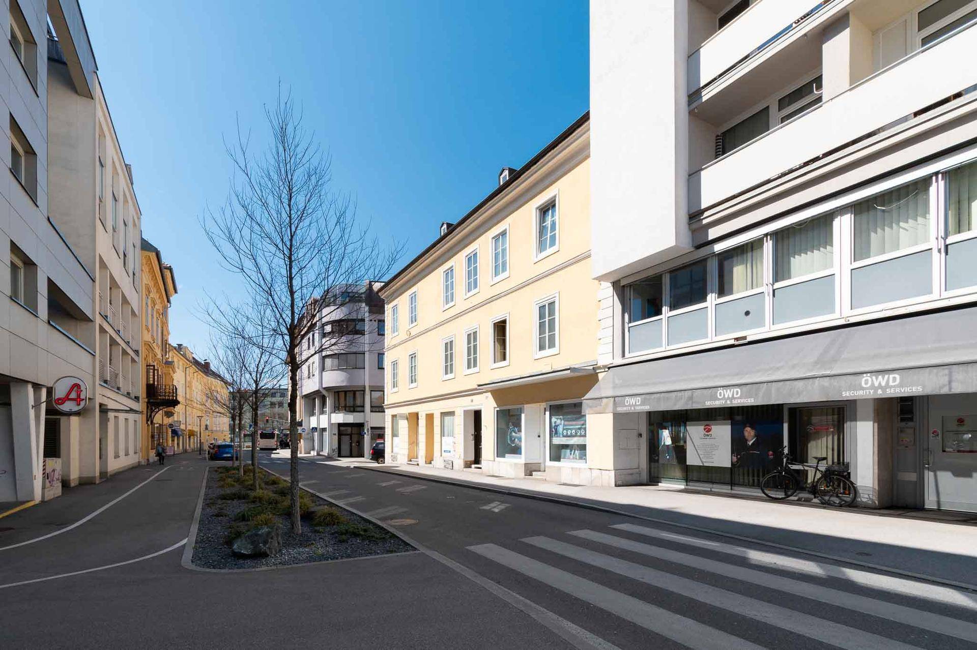 Belebte Straße mit Geschäften im Erdgeschoss und einem historischen Gebäude unter blauem Himmel.