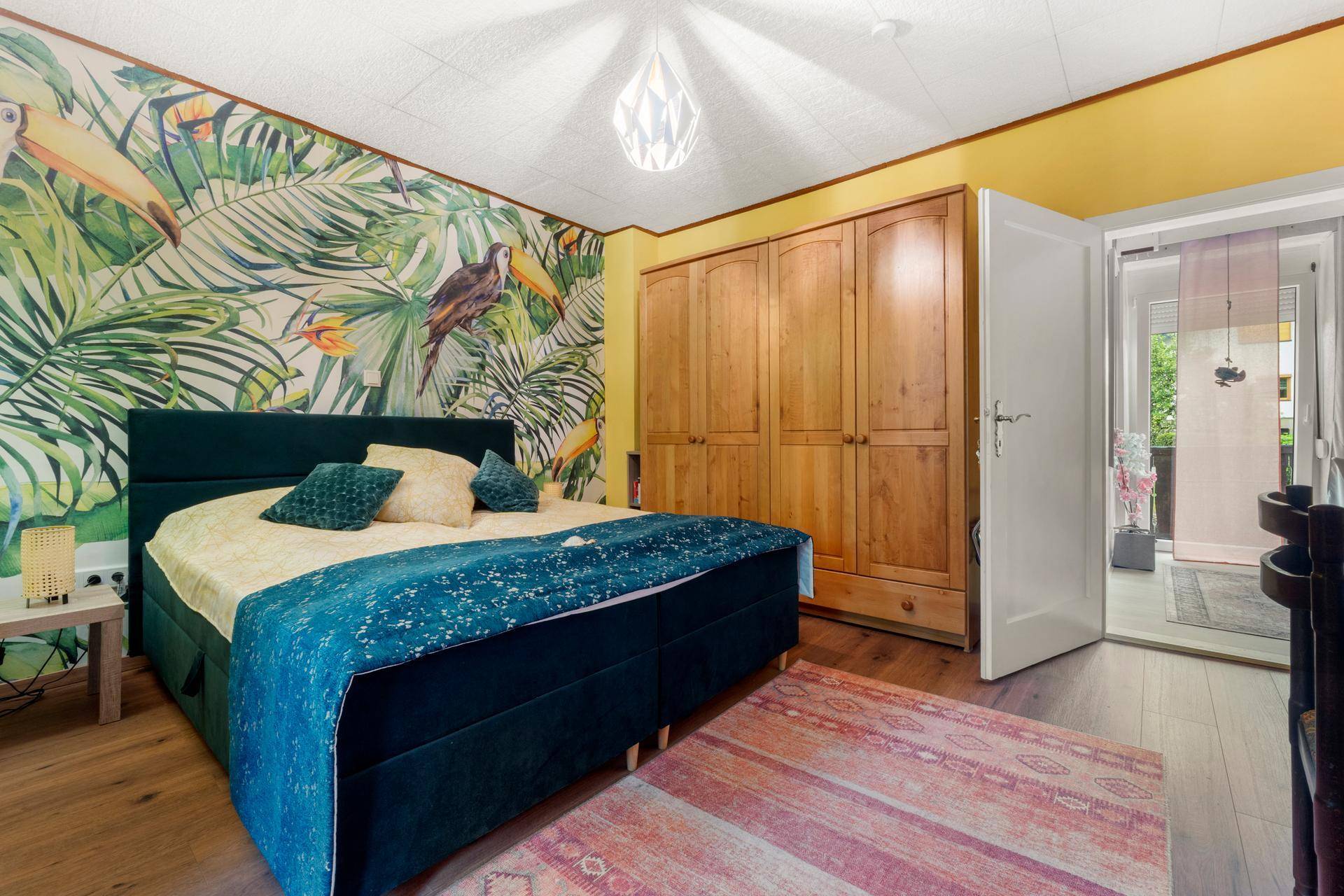 Farbenfrohes Schlafzimmer mit Doppelbett, Holzschrank und tropischer Tapete, Holzboden.
