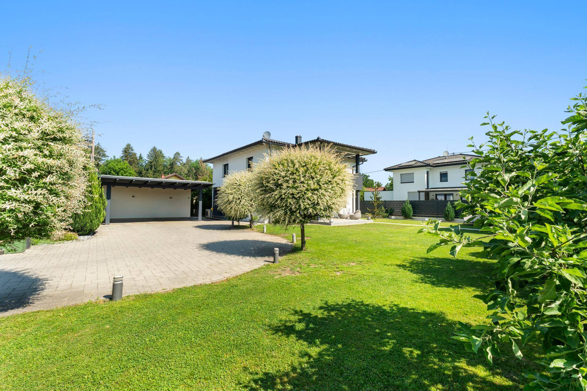 Großer gepflasterter Hof mit Carport und weitläufiger Rasenfläche, umgeben von Bäumen.