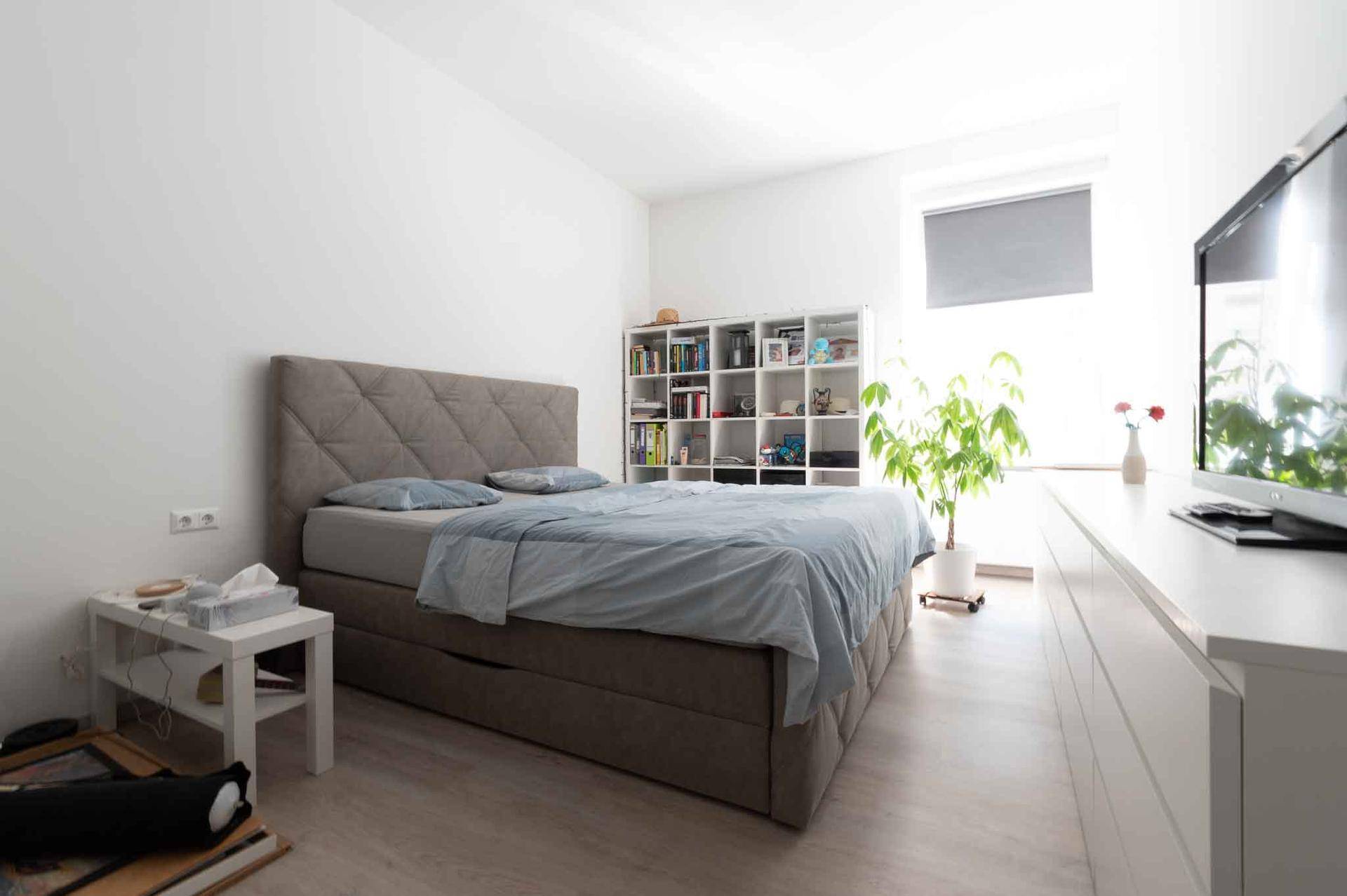 Helles Schlafzimmer mit großem Bett, weißem Regal und einem Fenster mit Rollo.