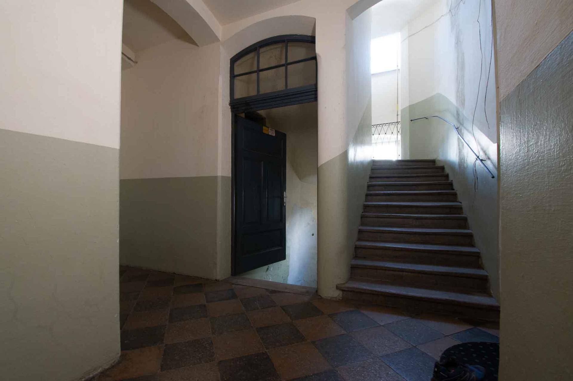 Historisches Treppenhaus mit gemusterten Fliesen, einer Holztür und einer geschwungenen Treppe.