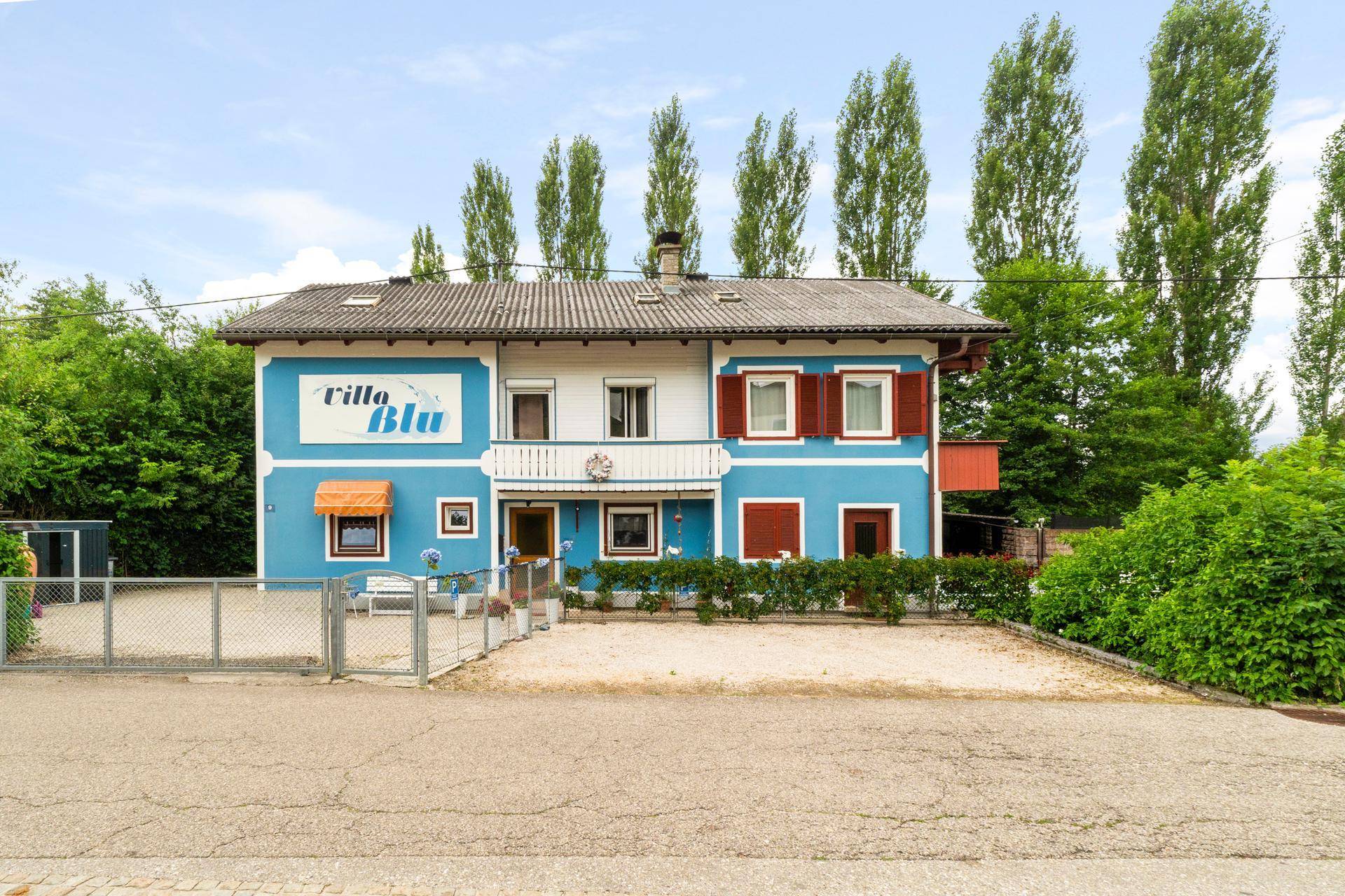 Vorderansicht des blauen Hauses mit weißer Fassade, umgeben von hohen Bäumen und einem Zaun.