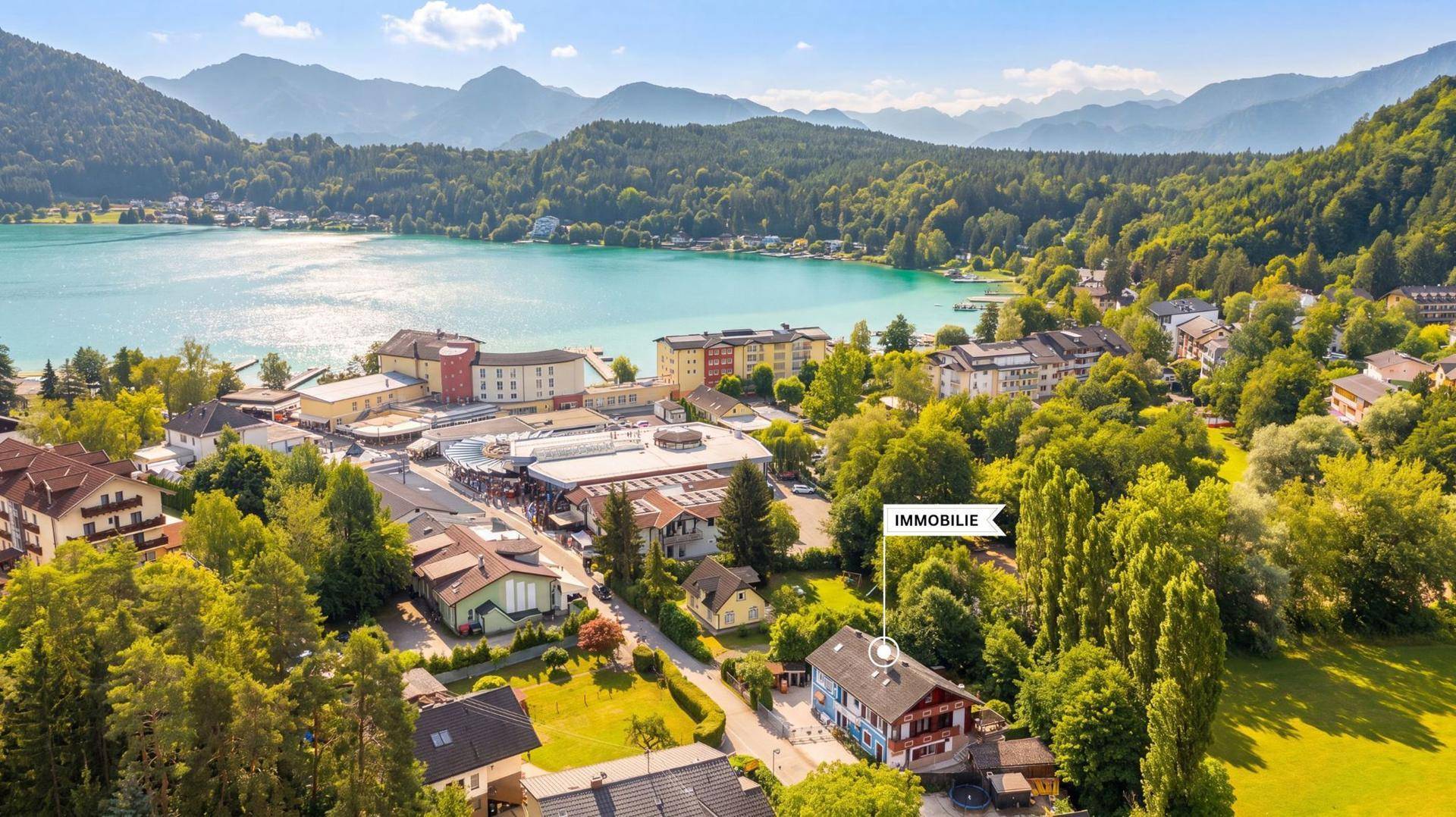 Panoramablick auf eine Siedlung am Seeufer mit Bergen im Hintergrund und markiertem Objekt.