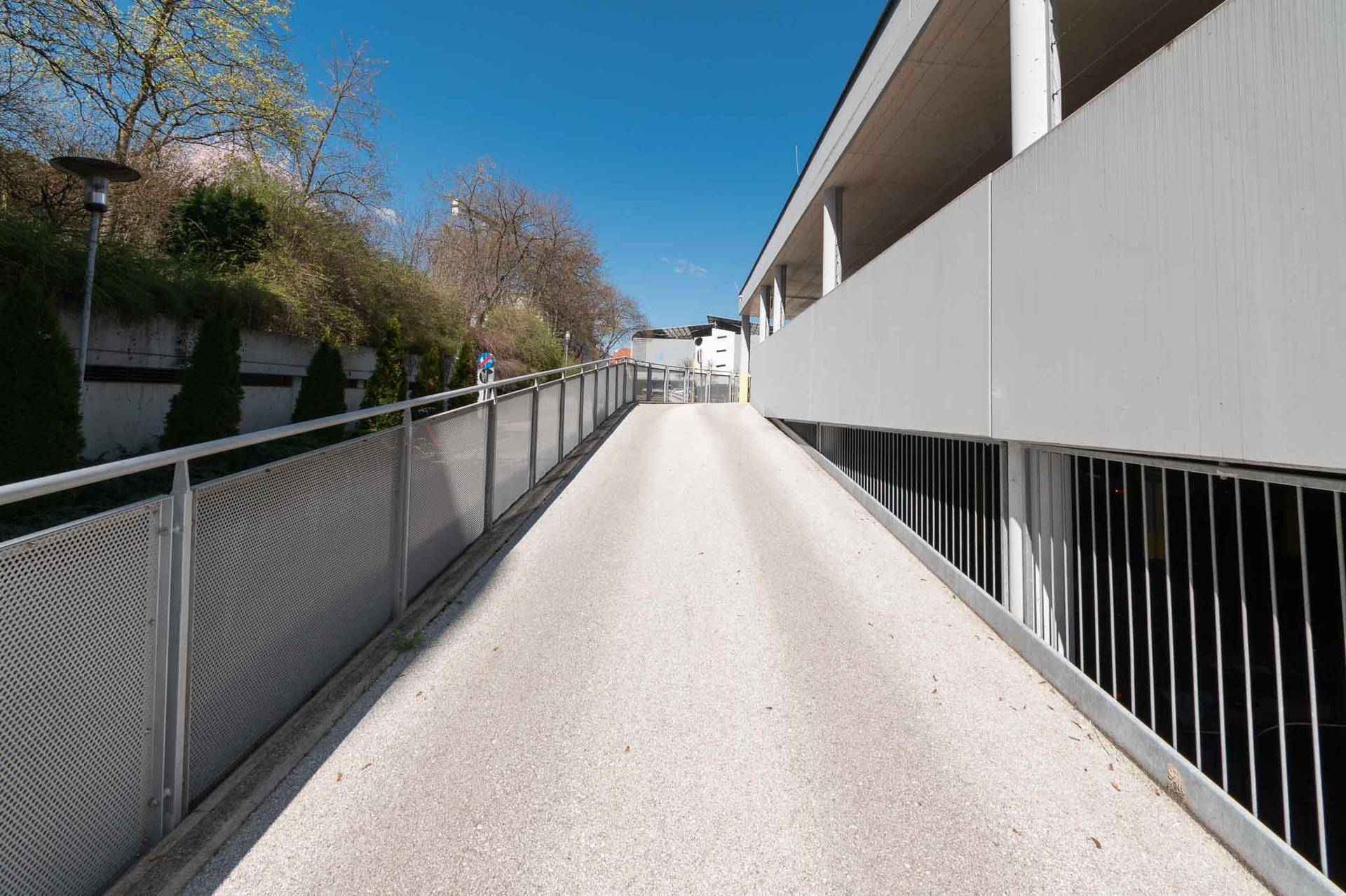 Betonrampe führt in ein Parkhaus, umgeben von Metallgeländern und Grünflächen.