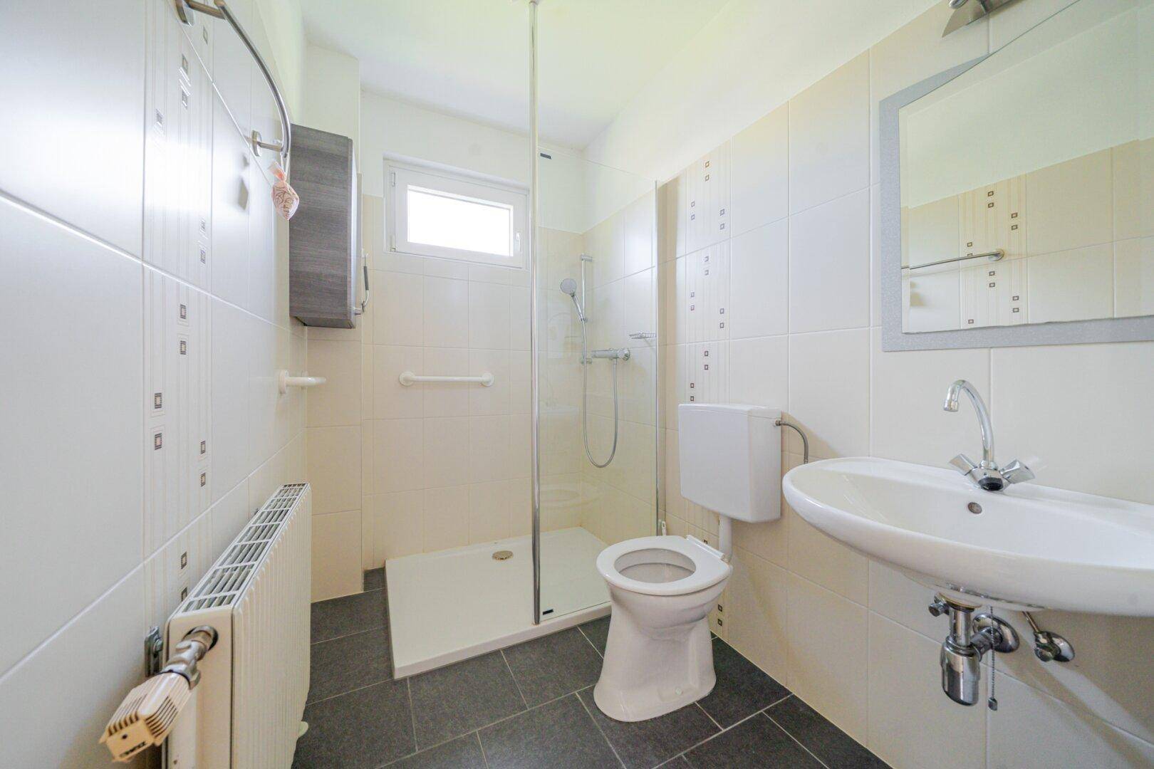 Helles Badezimmer mit Dusche, WC, Waschbecken und Fenster, ausgestattet mit weißen Fliesen.