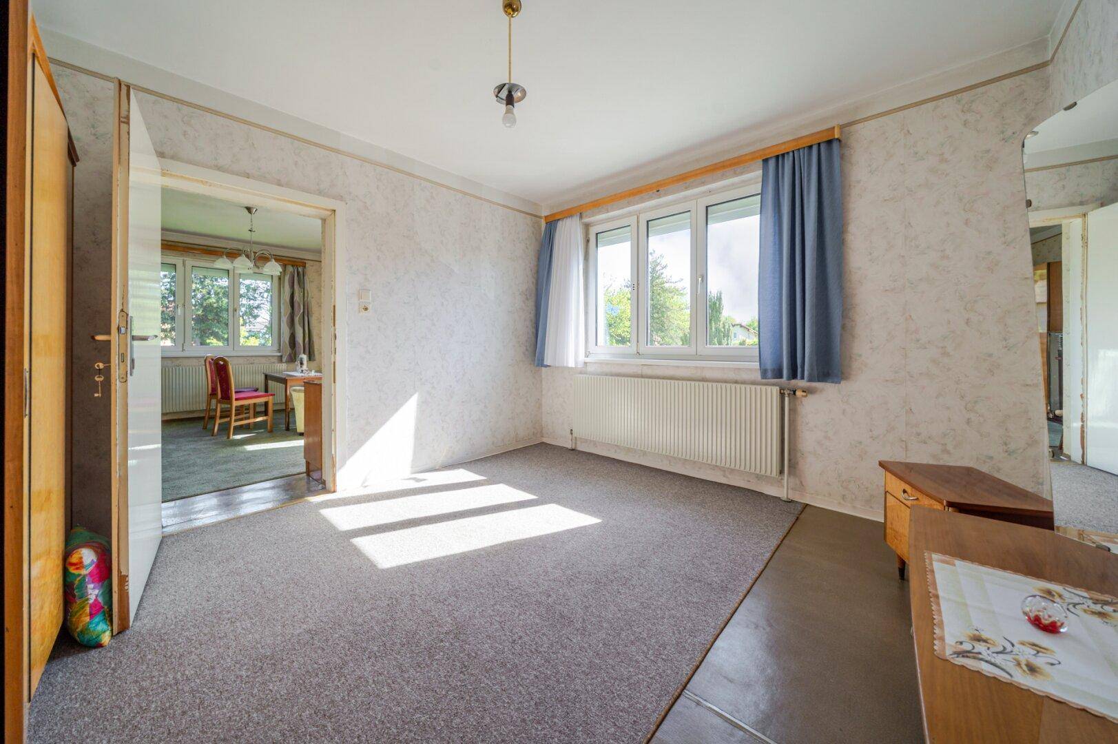 Helles Schlafzimmer mit Teppichboden, großem Fenster und Heizkörper unter blauem Vorhang.