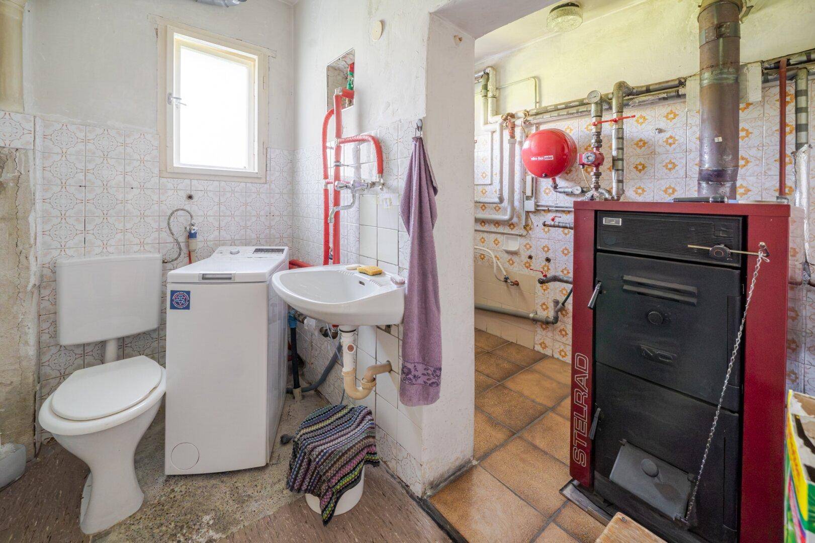 Hauswirtschaftsraum mit Heizungsanlage, Waschmaschine, WC und Waschbecken unter einem Fenster.