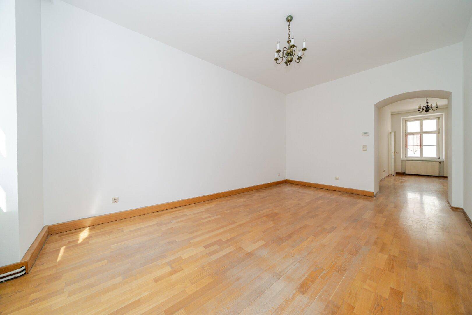 Geräumiges Zimmer mit Parkettboden, weiß gestrichenen Wänden und einem eleganten Kronleuchter.