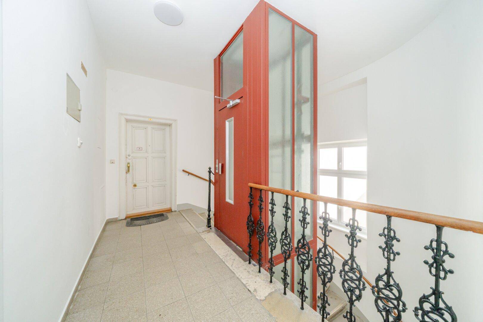 Treppenhaus mit modernem Aufzug, Fliesenboden und schmiedeeisernem Geländer im Gebäude.