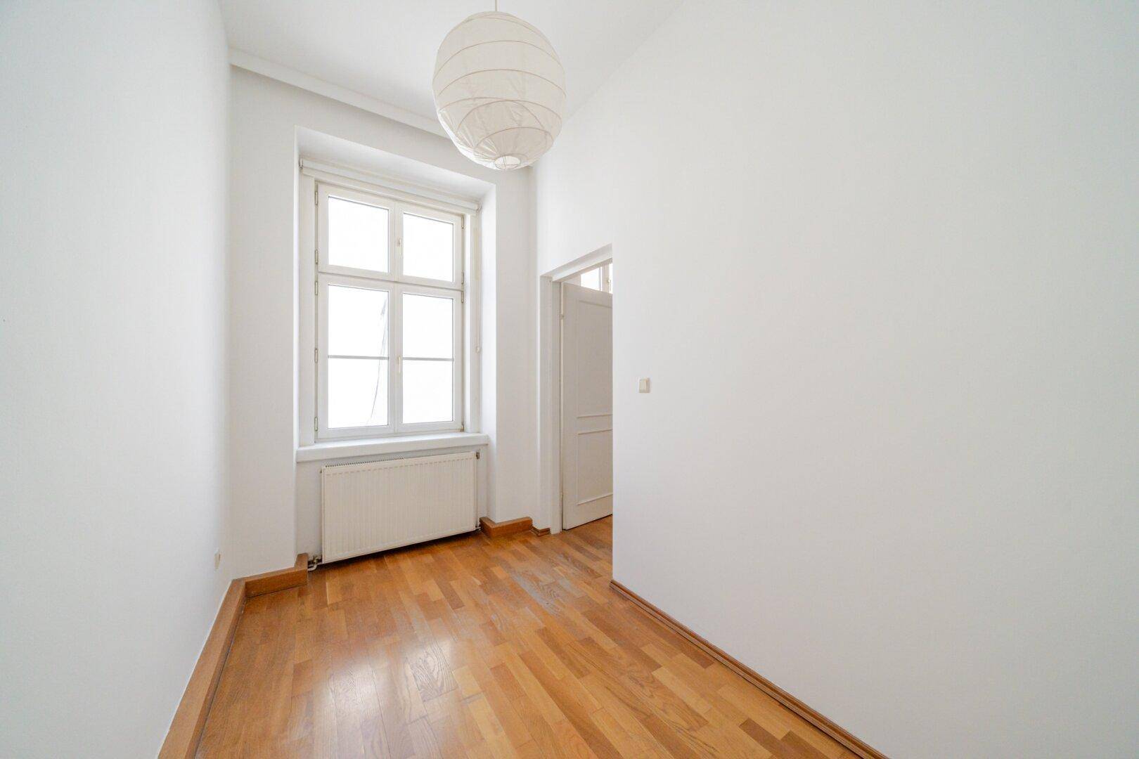 Kleineres Zimmer mit Parkettboden, weißer Deckenleuchte und Fenster mit Heizkörper darunter.