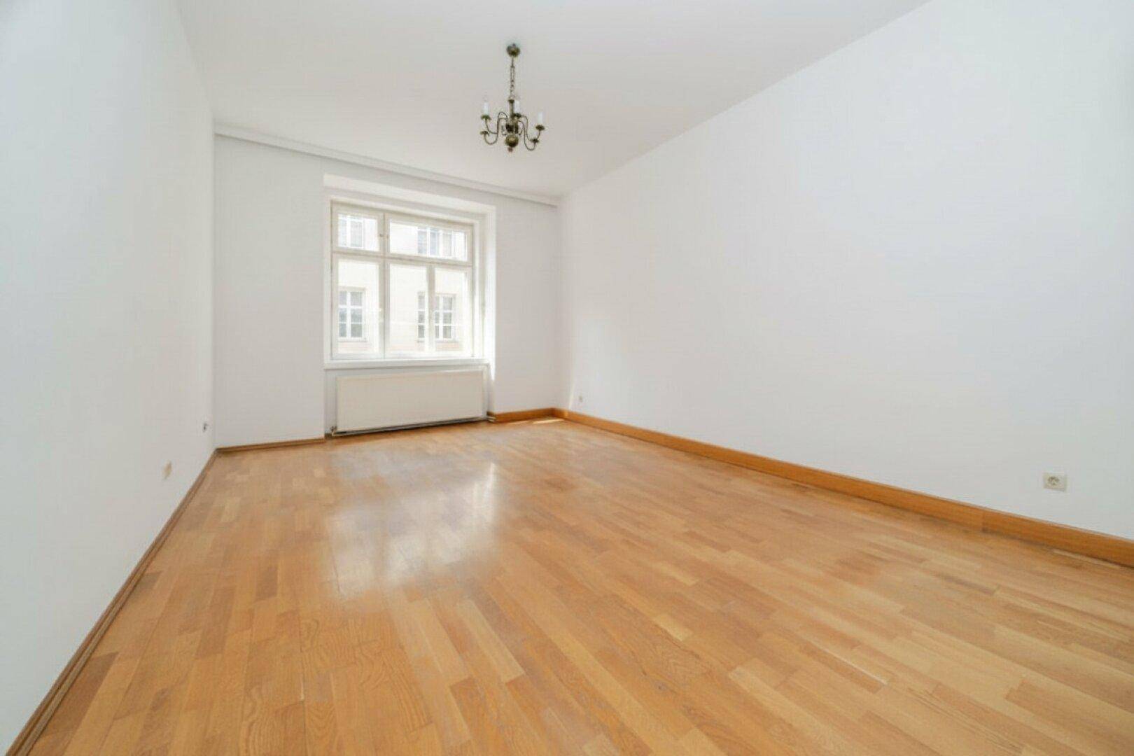 Helles Zimmer mit Parkettboden, weißen Wänden und klassischem Kronleuchter an der Decke.