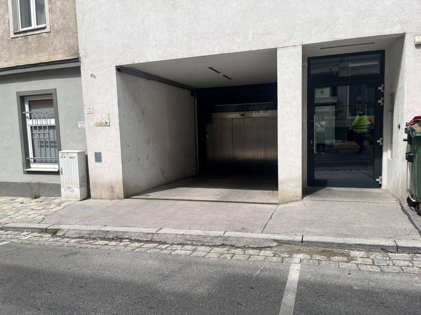 Eingangsbereich eines Gebäudes mit Zufahrt zu einer Tiefgarage und einer modernen Glastür.