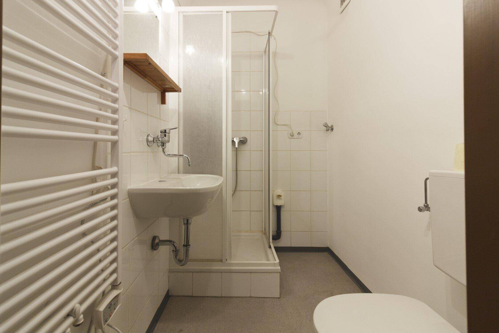 Kompaktes Badezimmer mit Dusche, Waschbecken, WC und beheiztem Handtuchhalter, hell gefliest.