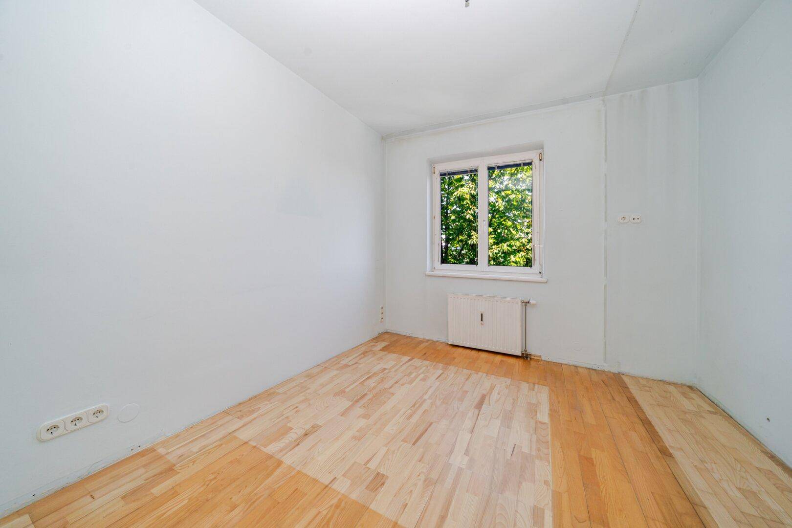 Leeres Schlafzimmer mit hellem Parkettboden, weißen Wänden und einem Fenster mit Blick ins Grüne.