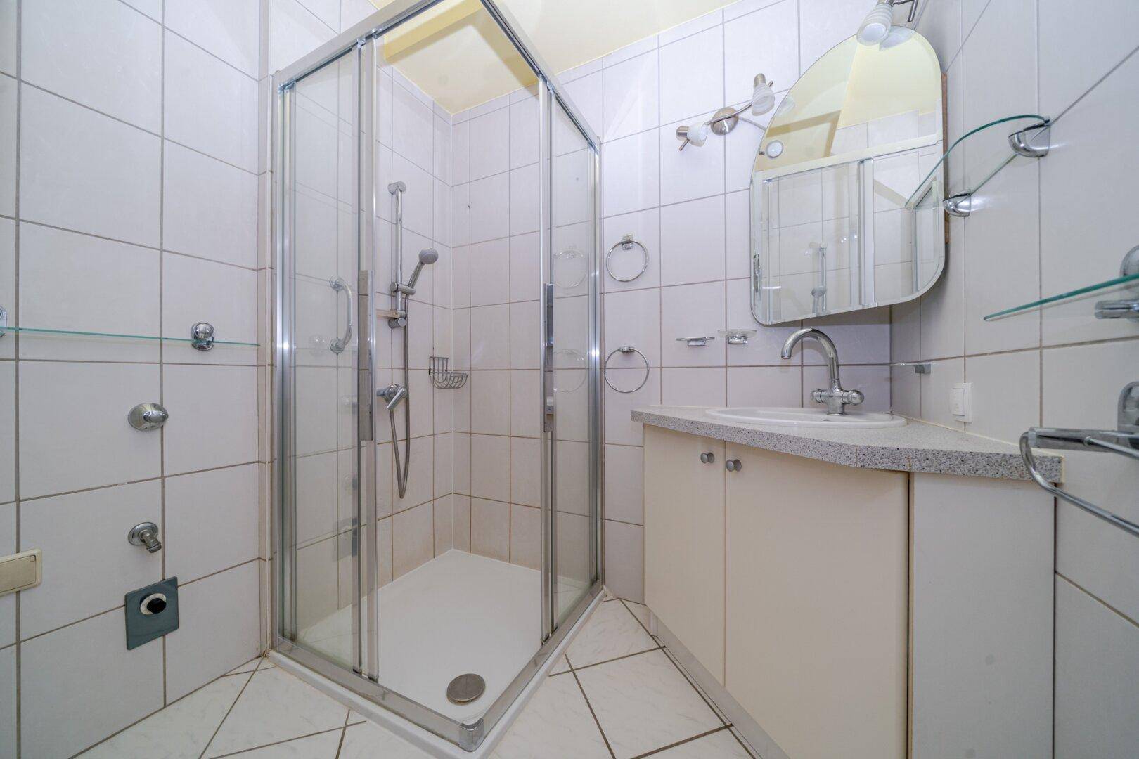 Kleines Badezimmer mit weißen Fliesen, einer Eckduschkabine und einem Waschbecken mit Spiegel.