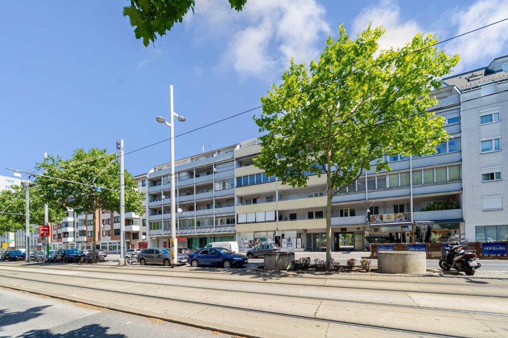 Straßenansicht eines Wohngebäudes mit Balkonen, parkenden Autos und Straßenbahnschienen, gesäumt von Bäumen.