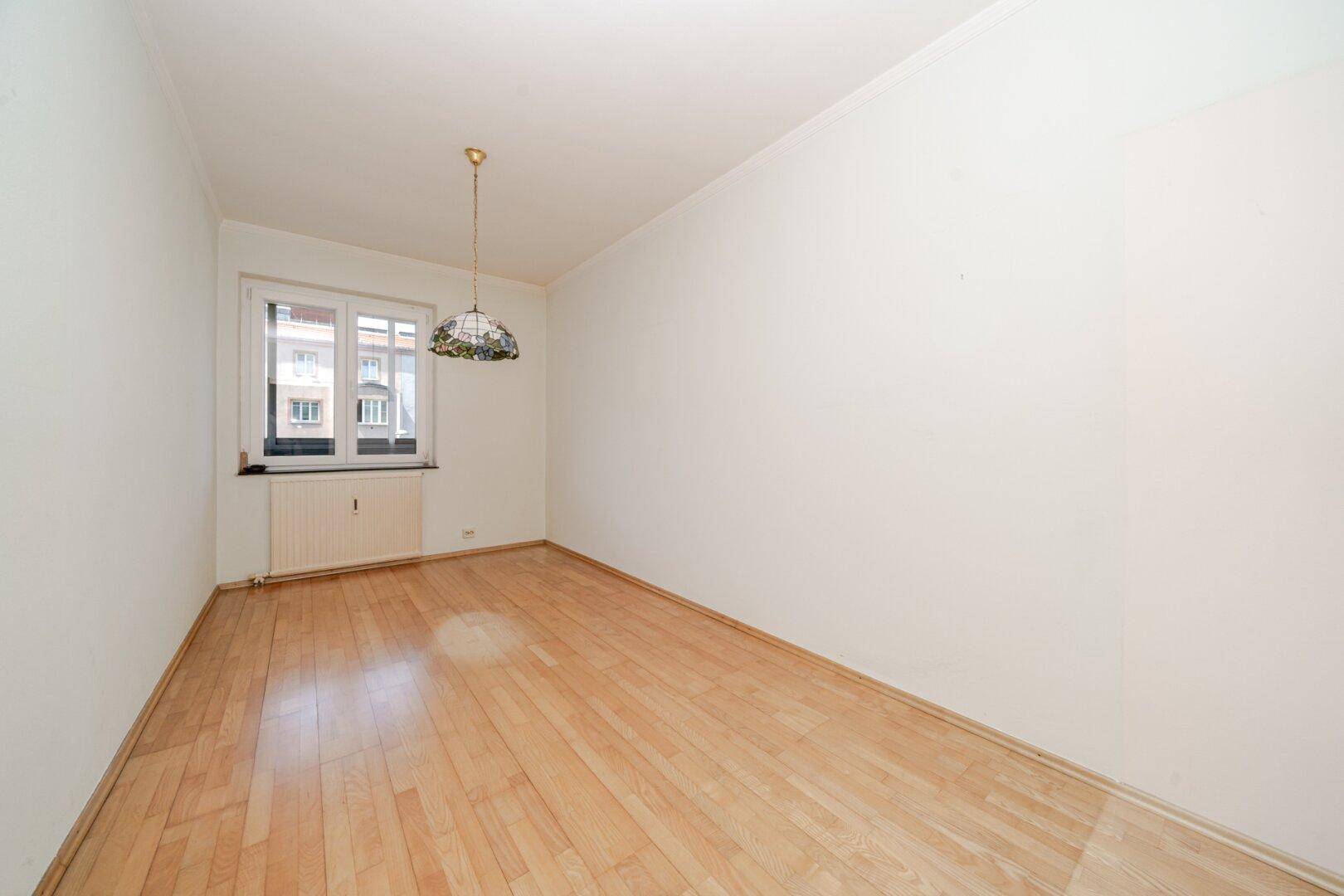 Helles, leeres Zimmer mit hellem Parkettboden, weißen Wänden und einem Fenster, ausgestattet mit einer Deckenlampe.