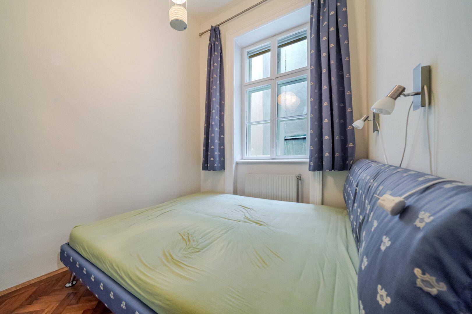 Schlafzimmer mit Doppelbett, Parkettboden und Fenster mit blauen Vorhängen.