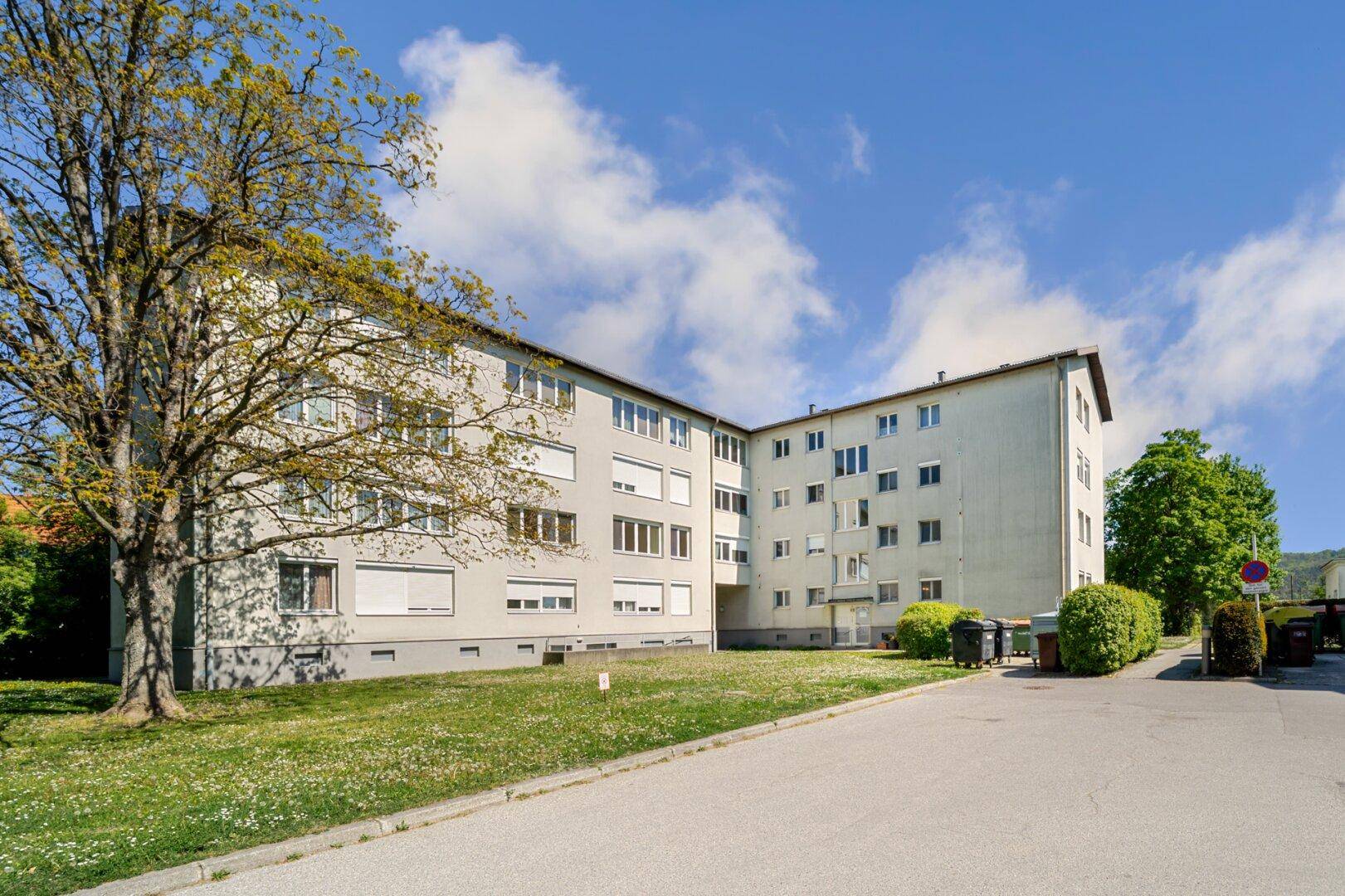Mehrstöckiges Wohngebäude mit grauer Fassade, Rollläden und gepflegter Grünanlage unter blauem Himmel.