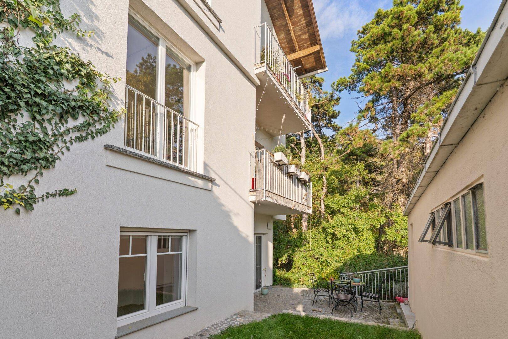 Außenansicht des Gebäudes mit Balkonen, einer kleinen Terrasse und üppigem Grün.