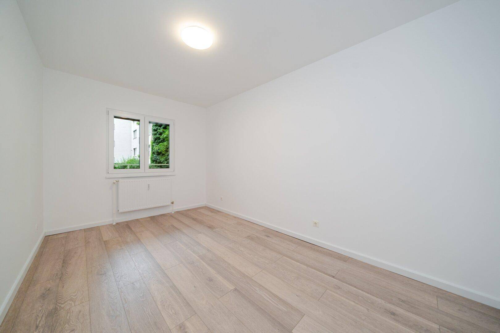Ruhiges Schlafzimmer mit hellem Laminatboden und einem Fenster mit Blick ins Grüne.