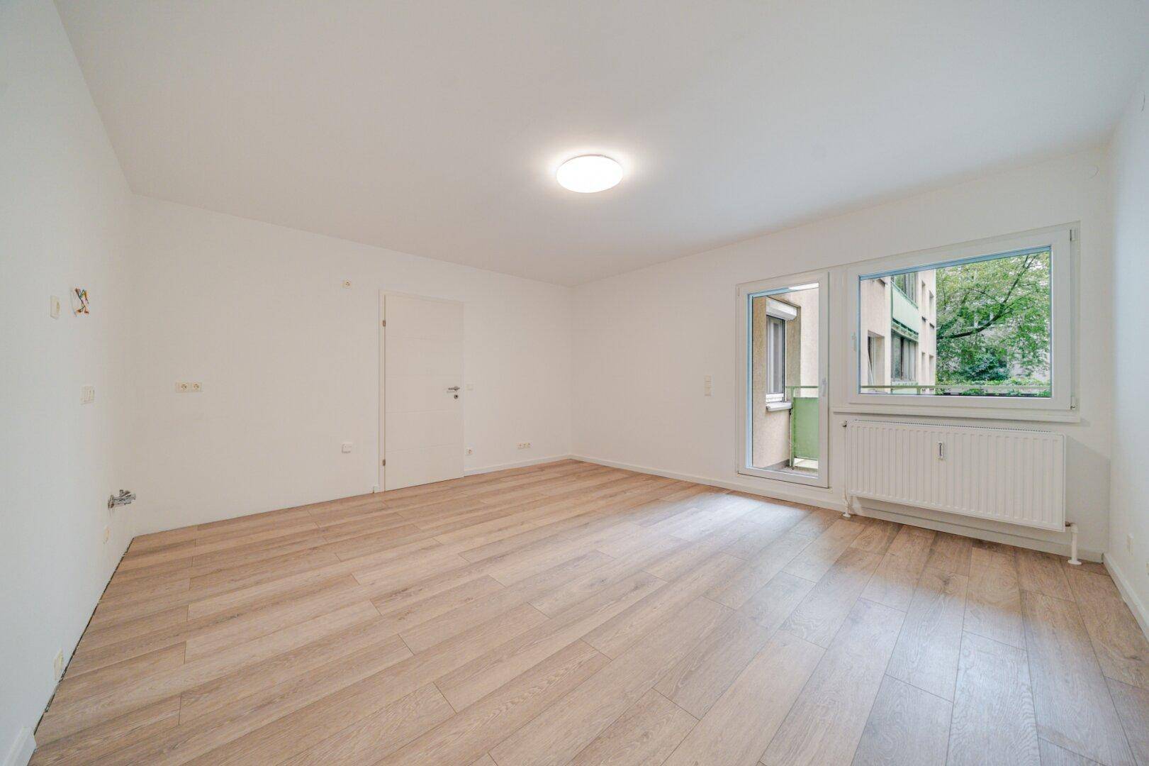 Helles, leeres Zimmer mit modernem Laminatboden und Zugang zum Balkon.