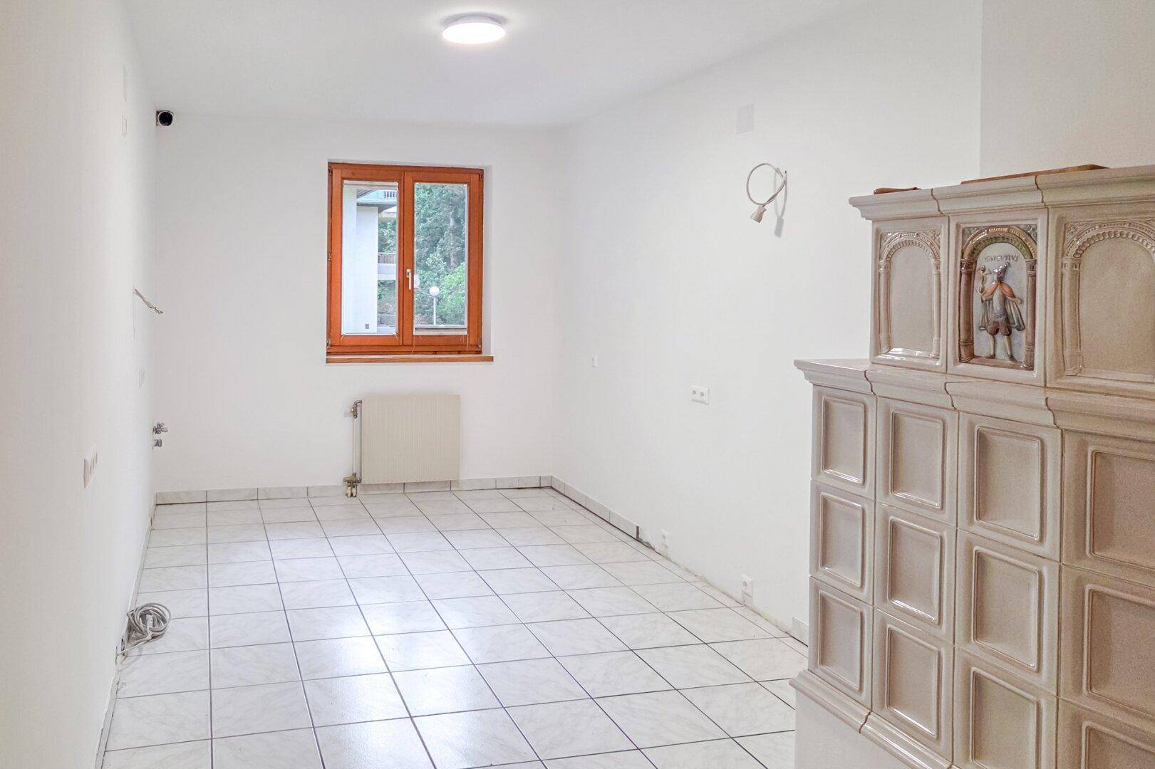 Heller, leerer Raum mit weiß gefliestem Boden, Holzfenster und einem traditionellen Kachelofen.