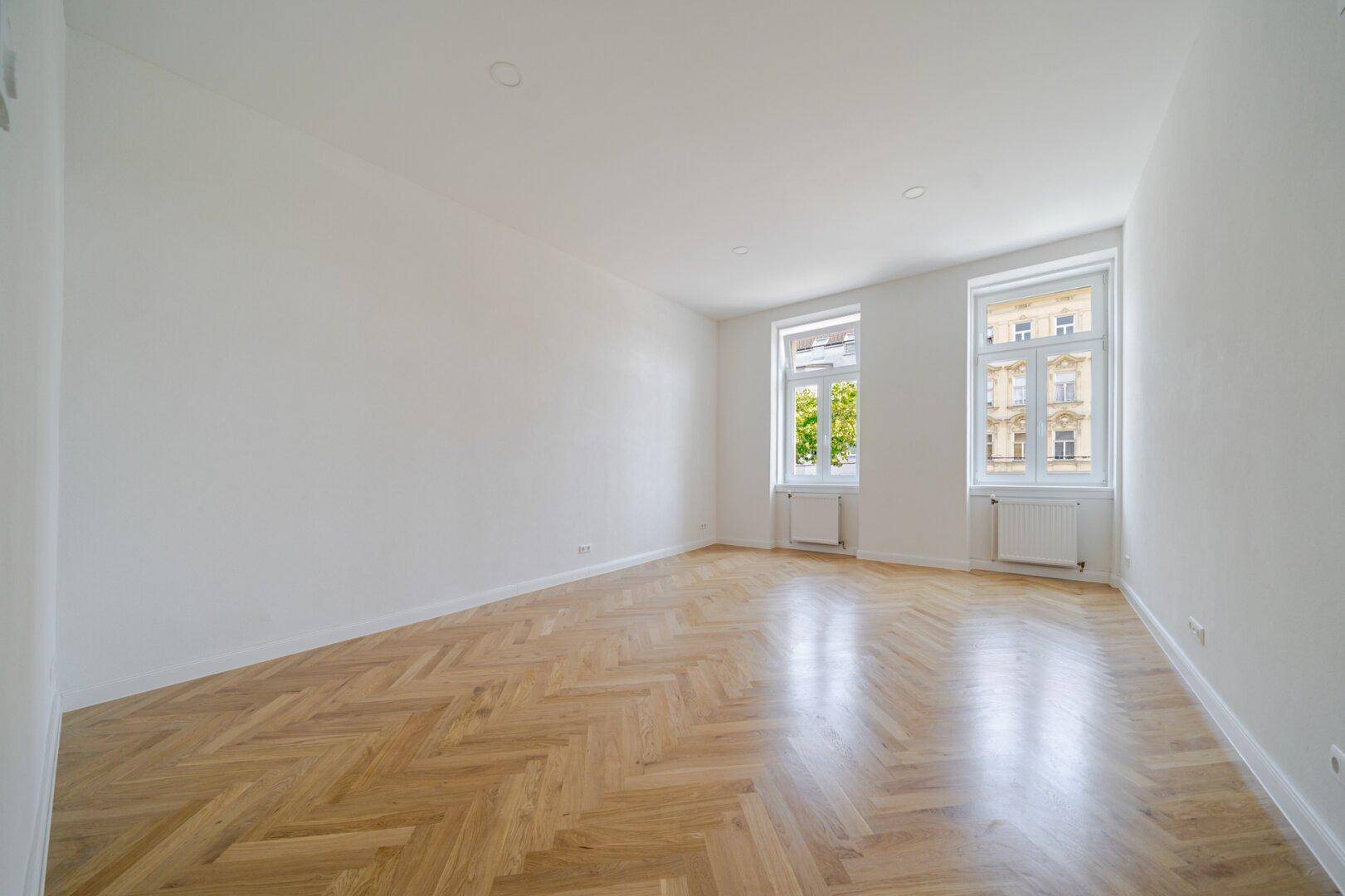 Geräumiges Zimmer mit glänzendem Fischgrätparkett und viel Tageslicht durch zwei Fenster.