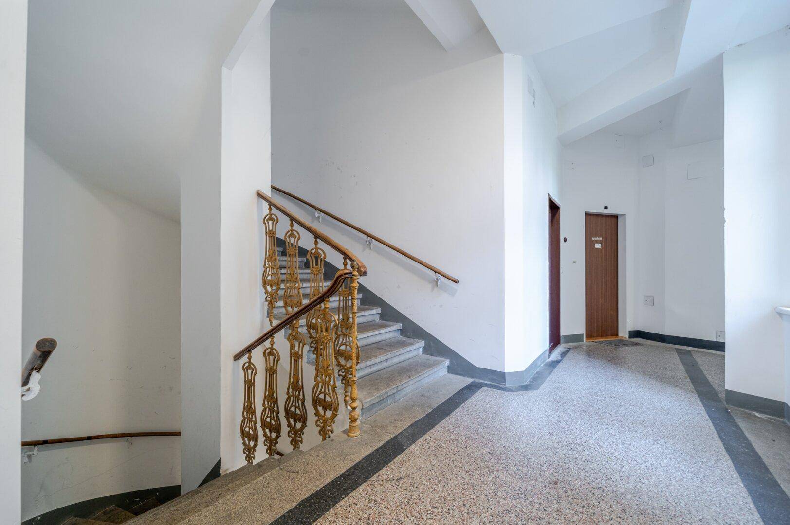 Historisches Treppenhaus mit kunstvollen Geländern, Terrazzoboden und Zugang zum Aufzug.
