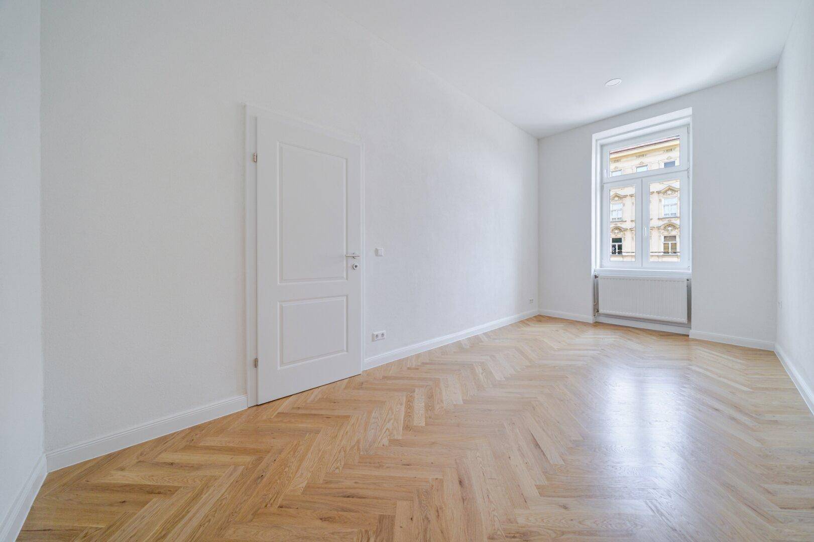 Ruhiges Zimmer mit klassischem Fischgrätparkett, weißen Wänden und einem großen Fenster.