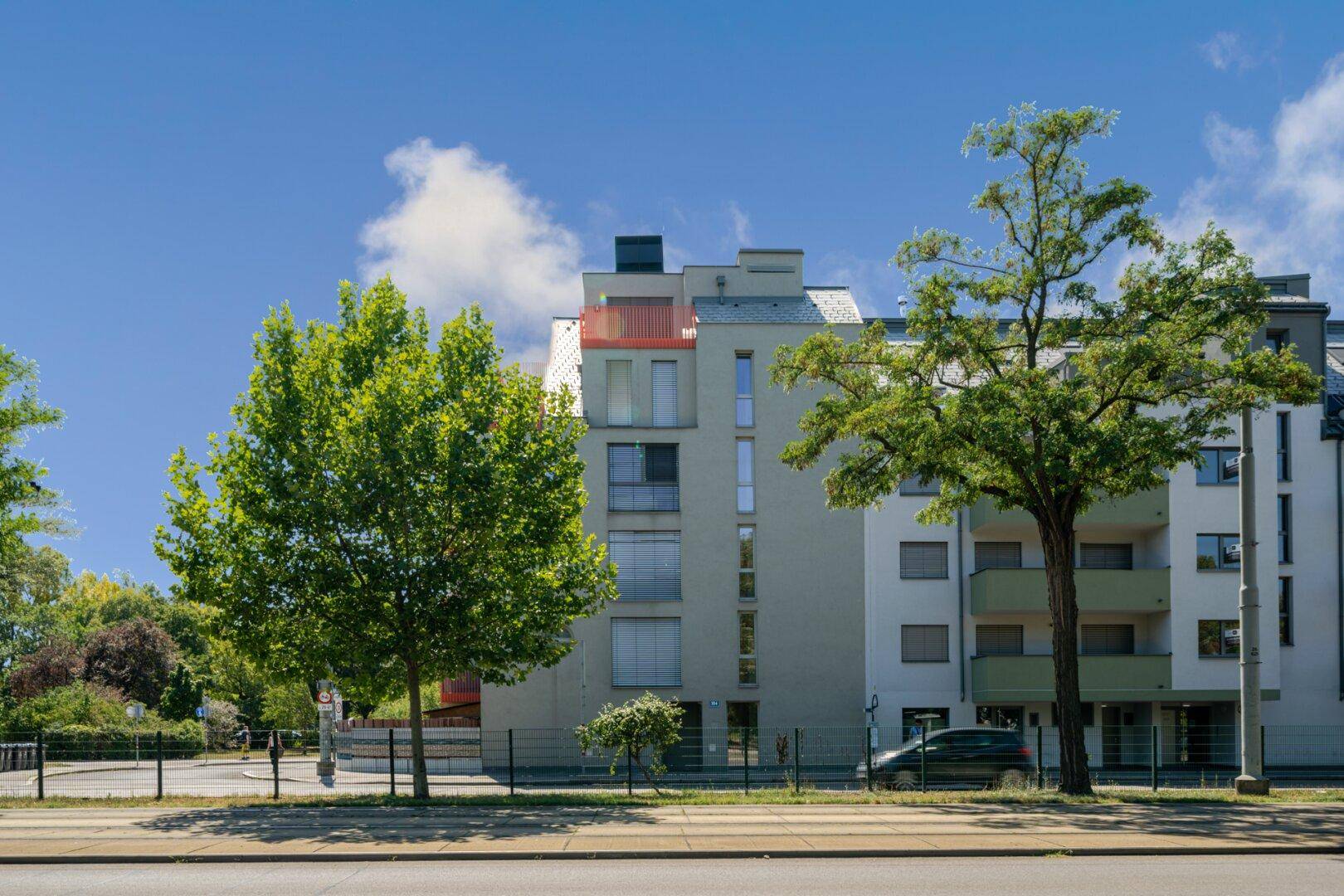 Moderne Wohngebäude-Fassade mit großen Fenstern, Rollläden und grüner Umgebung an einem sonnigen Tag.