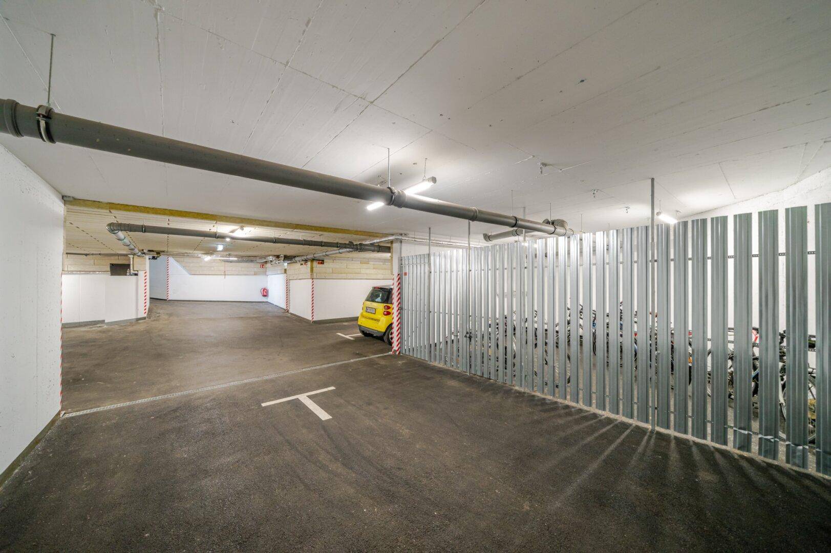 Tiefgarage mit markierten Stellplätzen, Fahrradabstellbereich und sichtbaren Installationen an der Decke.