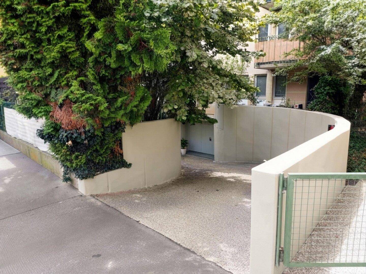 Geschwungene Einfahrt zu einer Tiefgarage, umgeben von üppigem Grün und einem Zaun.