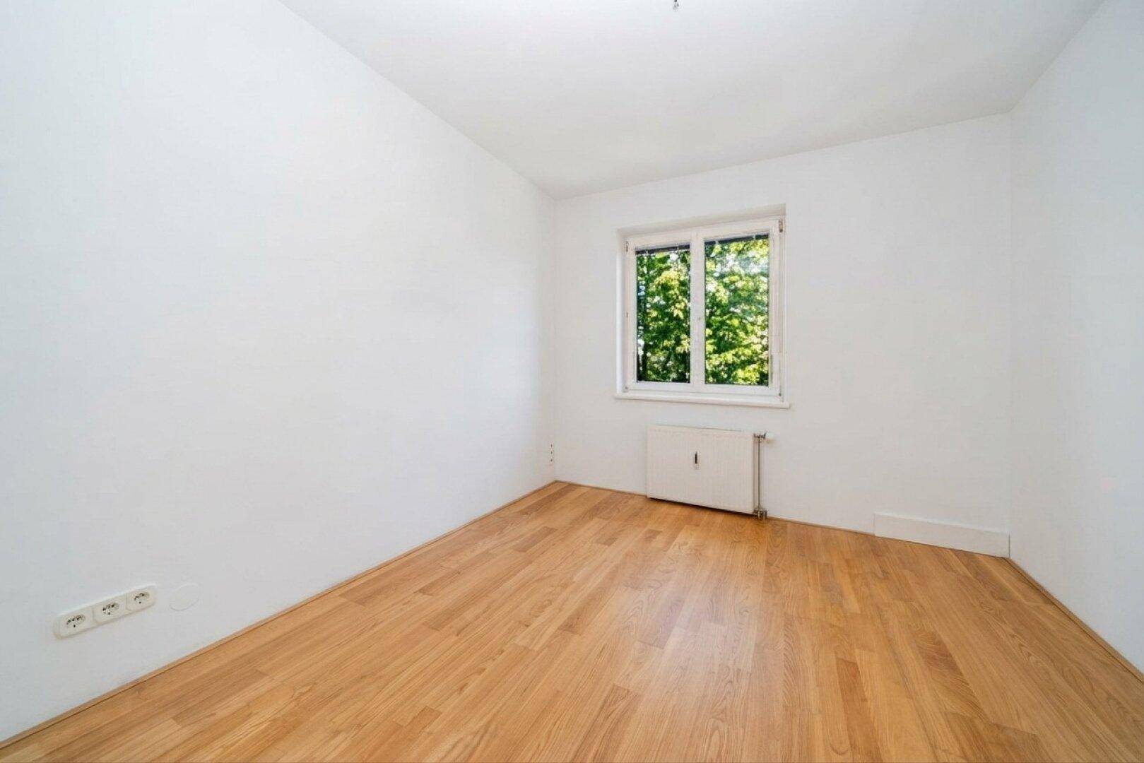 Leeres Zimmer mit hellem Holzboden, weißen Wänden und Fenster mit Blick ins Grüne.