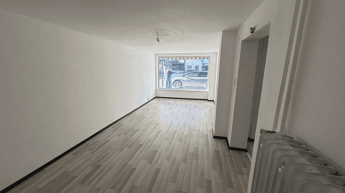 Heller, leerer Raum mit großen Fenstern zur Straße, hellem Laminatboden und einem Heizkörper.
