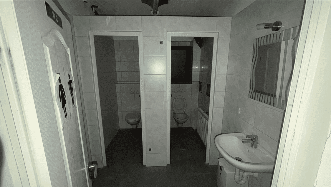 Öffentlicher Toilettenbereich mit zwei Kabinen, einem Waschbecken und gefliesten Wänden und Böden.