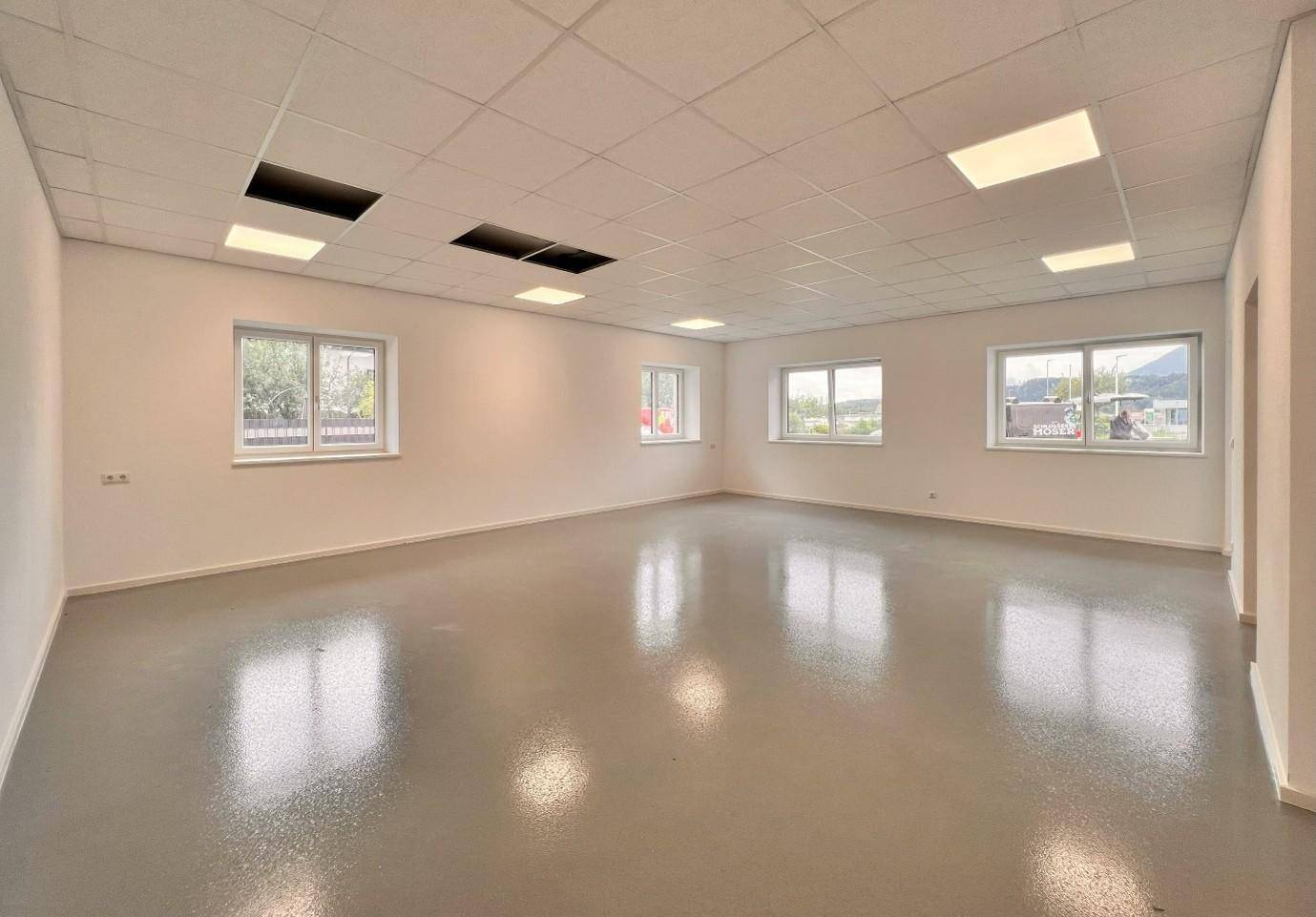 Geräumiger, leerer Gewerberaum mit mehreren Fenstern, hellem Boden und einer Rasterdecke.