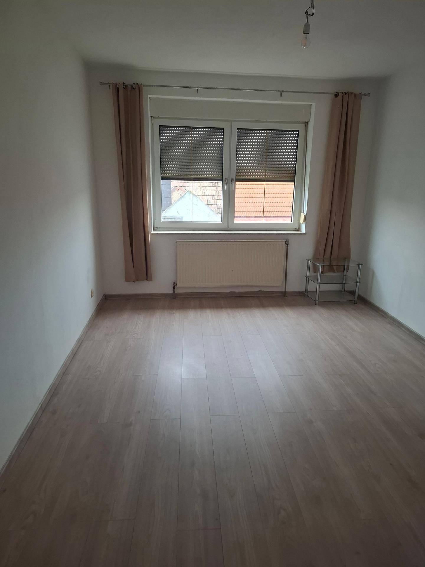 Leeres Schlafzimmer mit Laminatboden, großem Fenster und Heizkörper unter dem Fenster.