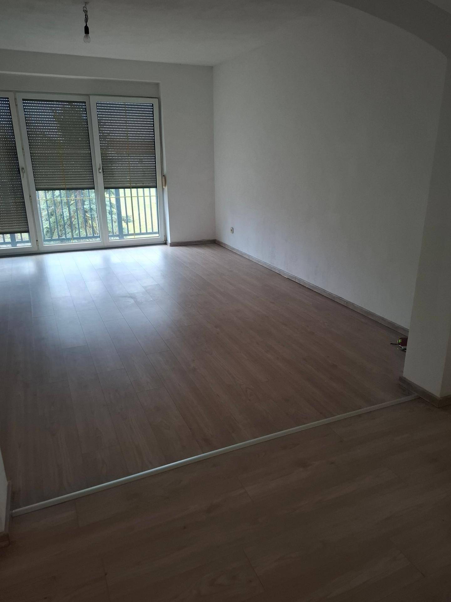 Geräumiges Wohnzimmer mit Laminatboden und großen Fenstern mit Rollläden.