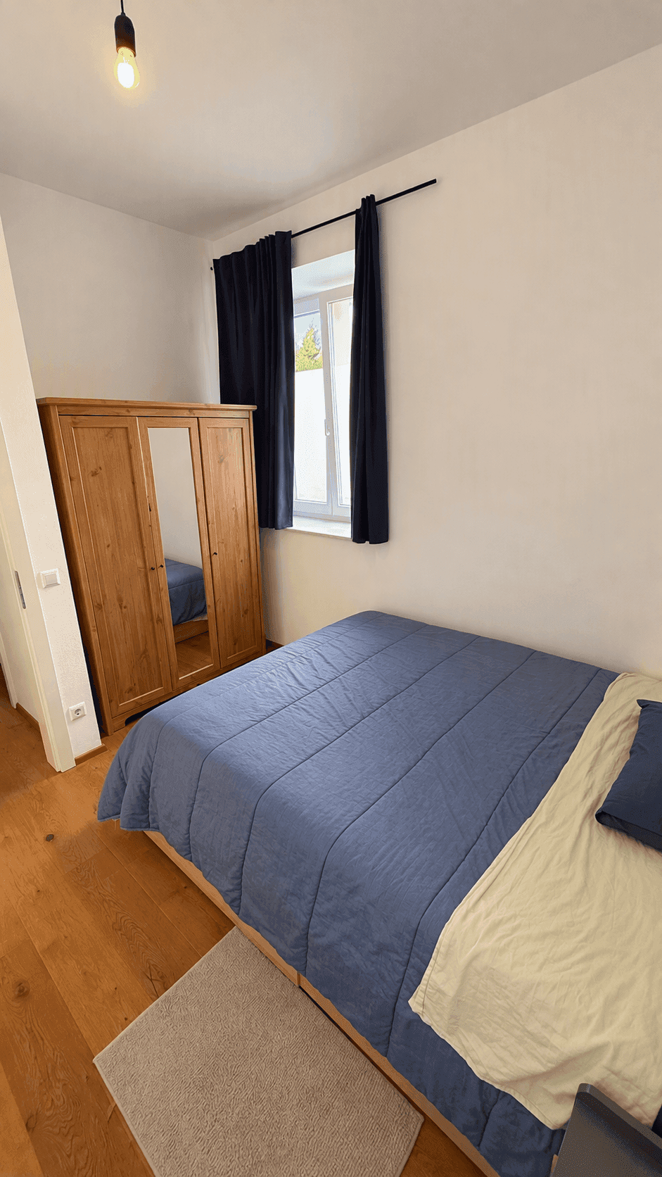 Schlafzimmer mit Doppelbett, Holzschrank und Fenster mit dunklen Vorhängen.