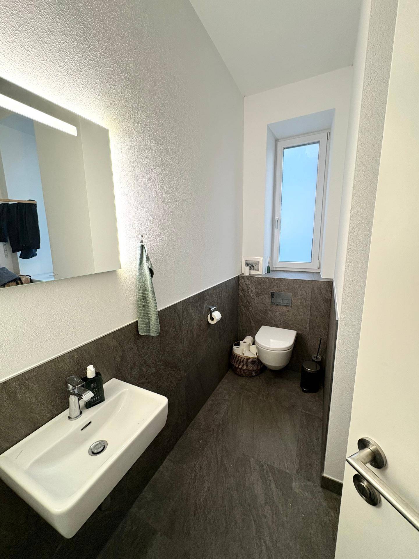Modernes Gäste-WC mit dunklen Fliesen, wandhängendem WC und kleinem Fenster.