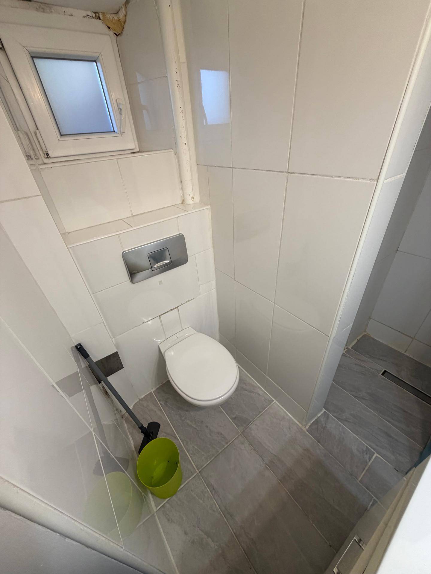 Toilette mit Wandspülkasten und einem kleinen Fenster, umgeben von hellen Fliesen.