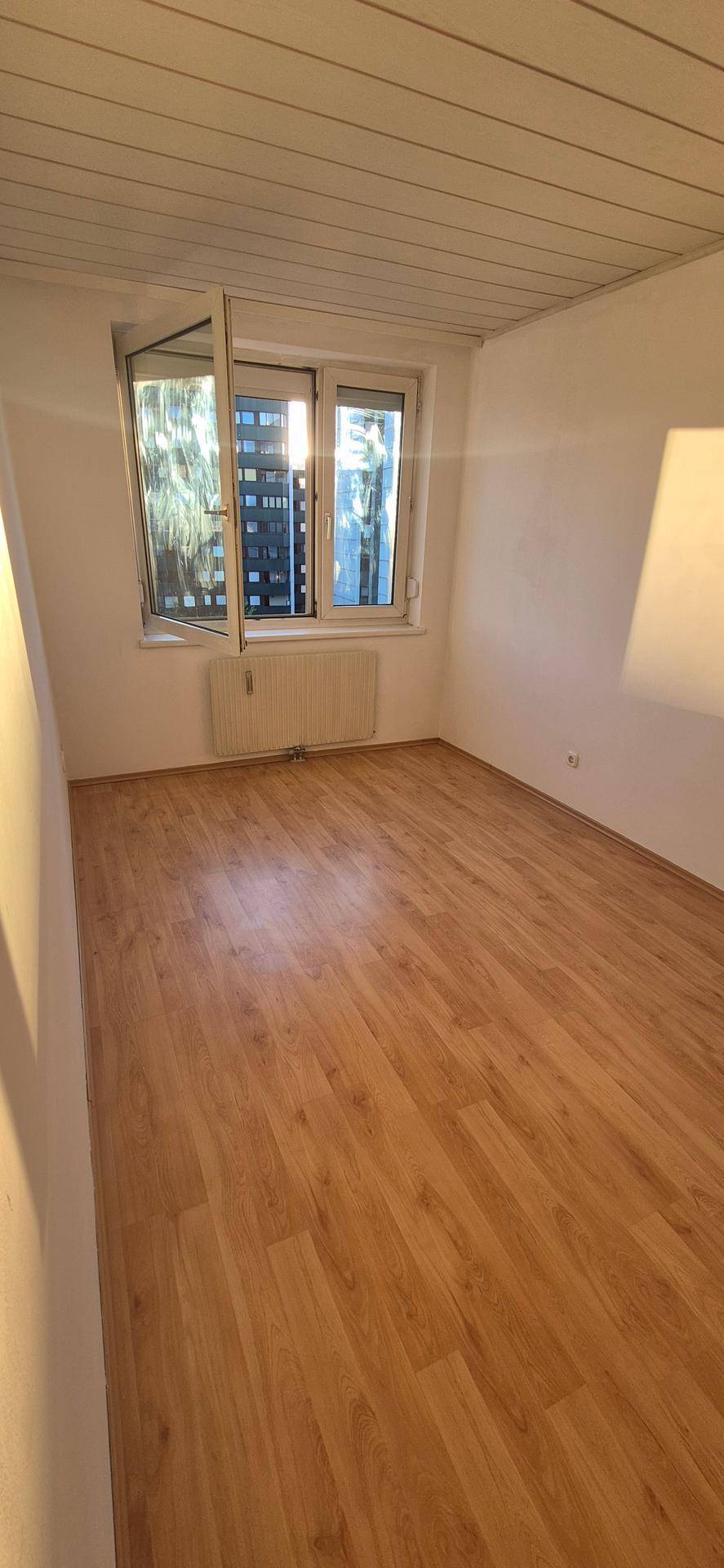 Helles Zimmer mit Laminatboden, Holzdecke und einem Fenster mit Blick auf ein Gebäude.
