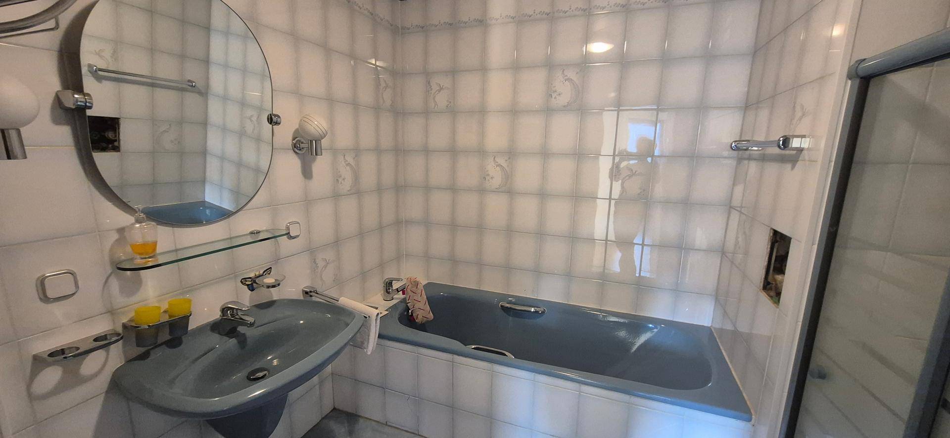 Badezimmer mit hellblauen Fliesen, passender Badewanne und Waschbecken sowie einem runden Spiegel.