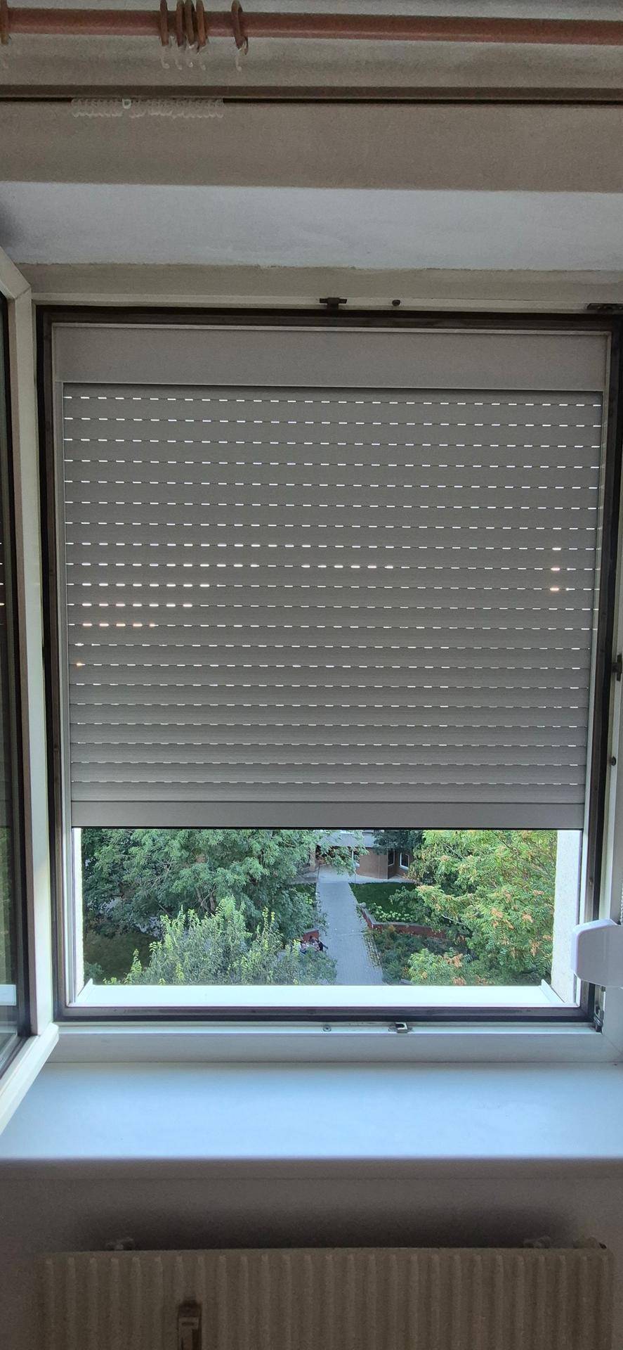 Fenster mit geschlossenem Rollladen und Blick auf grüne Bäume im Außenbereich.