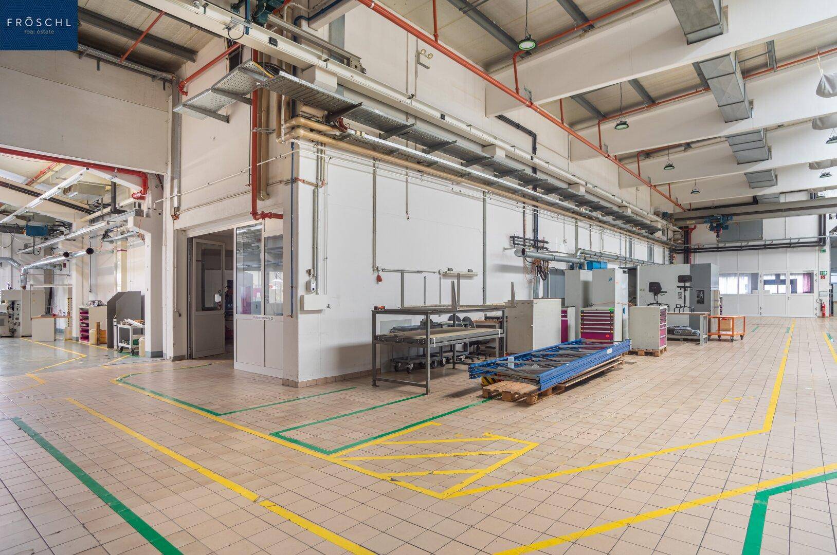 Produktionshalle mit Fliesenboden, gelben Markierungen und einem abgetrennten Bürobereich im Hintergrund.