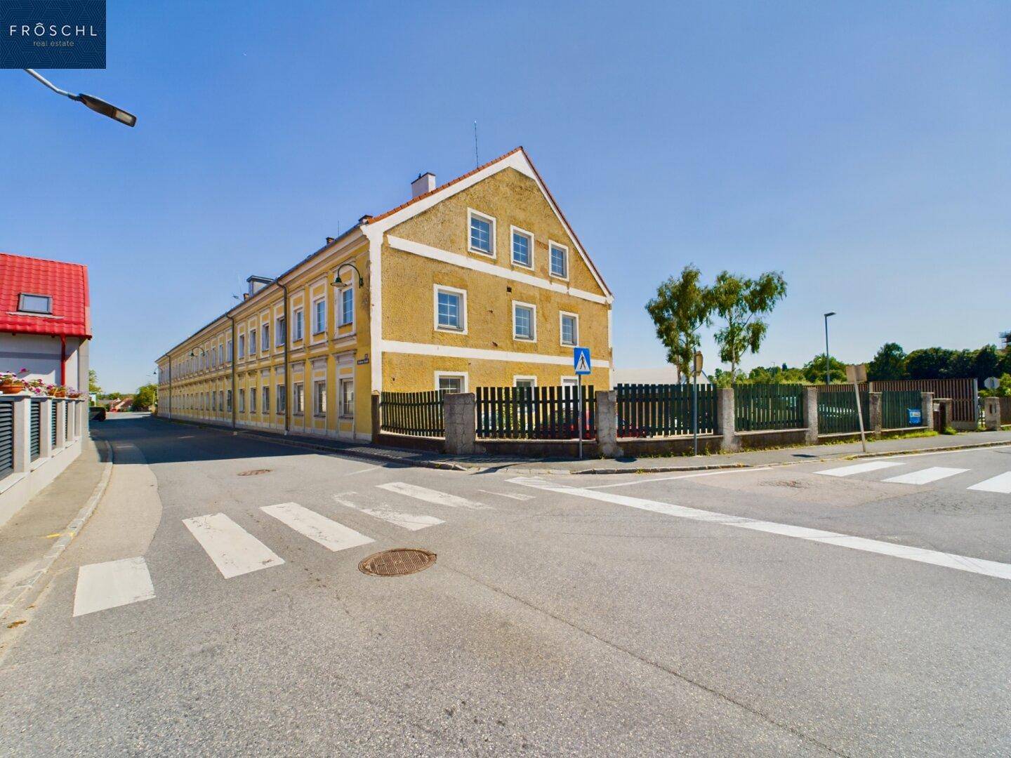 Historisches Gebäude mit gelber Fassade an einer Straßenecke, umgeben von einem Zaun und Bäumen.
