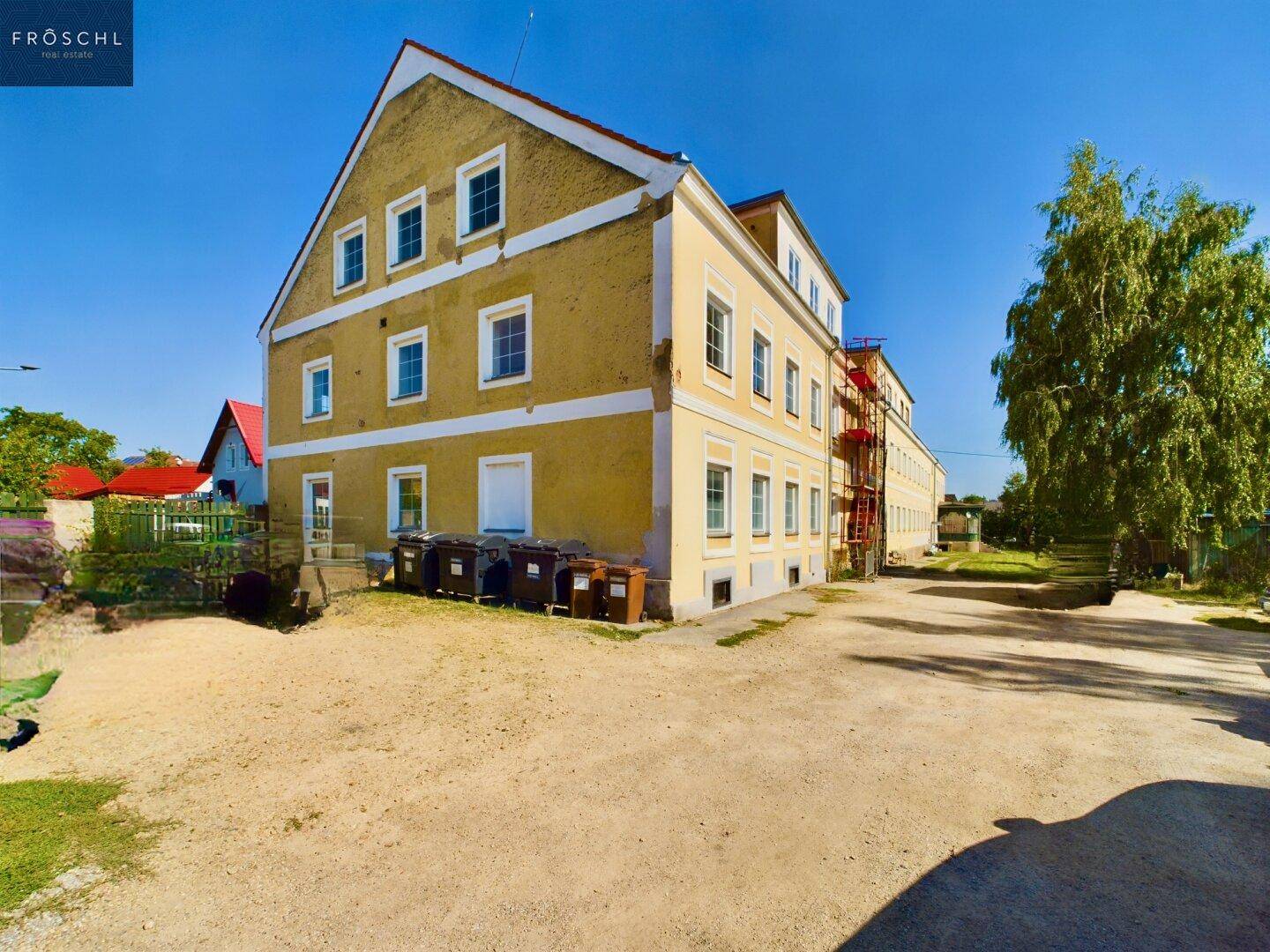 Seitenansicht des historischen Gebäudes mit gelber Fassade, einem Schotterparkplatz und Mülltonnen.