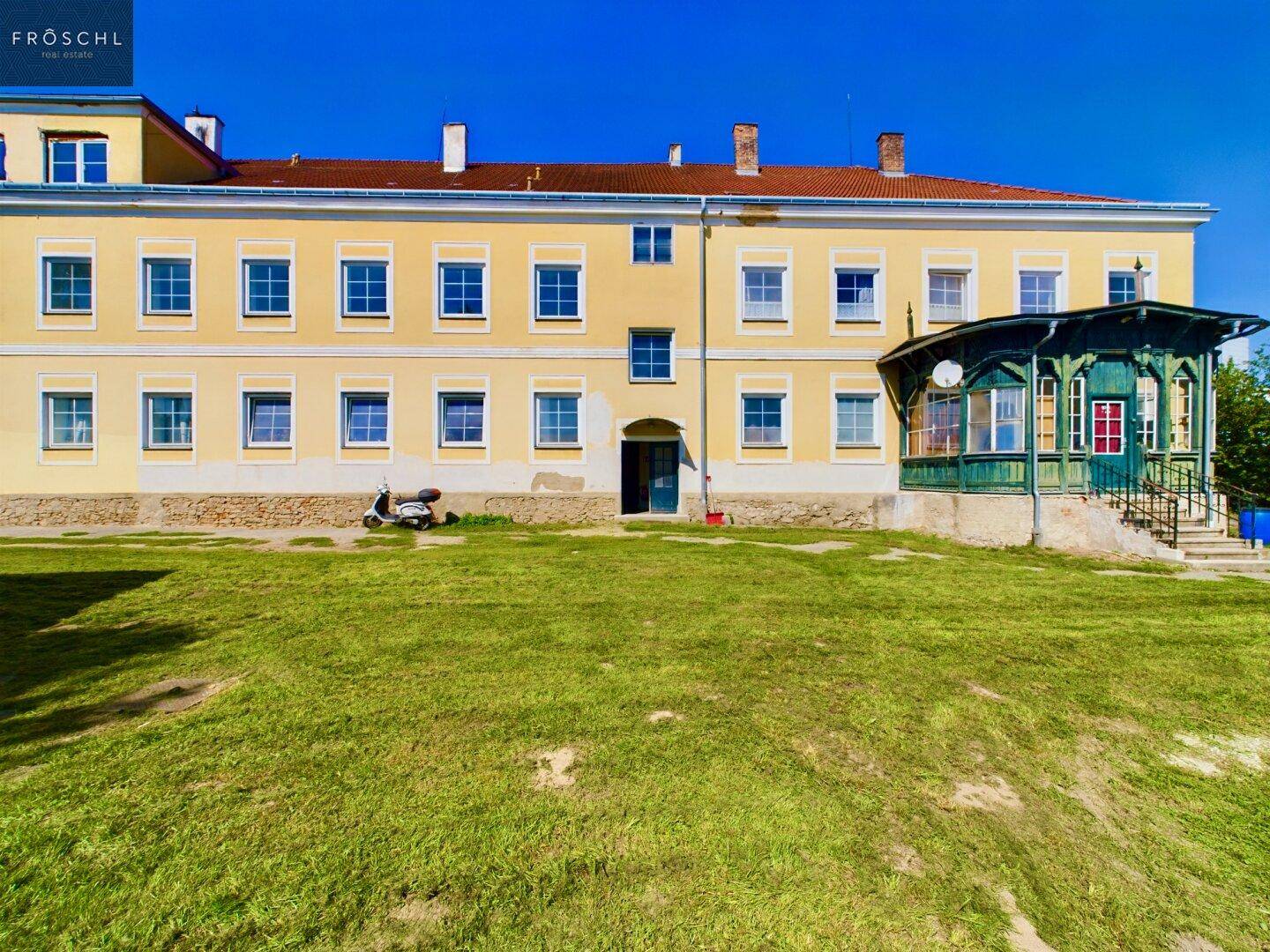 Ansicht des historischen Gebäudes mit gelber Fassade, vielen Fenstern und einem großen Rasenbereich davor.
