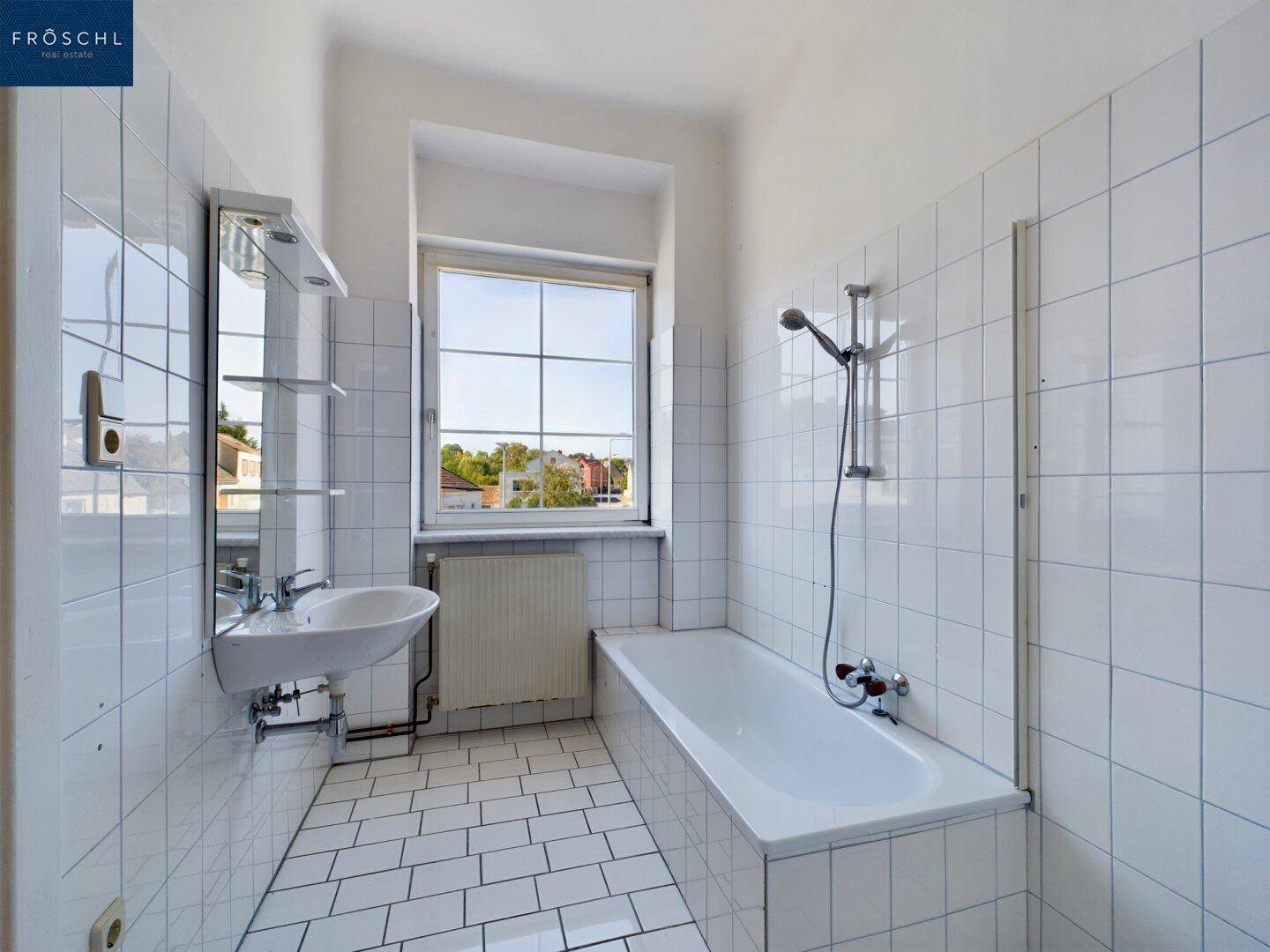 Helles Badezimmer mit weißen Fliesen, Badewanne, Waschbecken und einem Fenster.